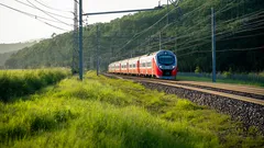 Moderní červený vlak projíždí krajinou se zelení a elektrickým vedením